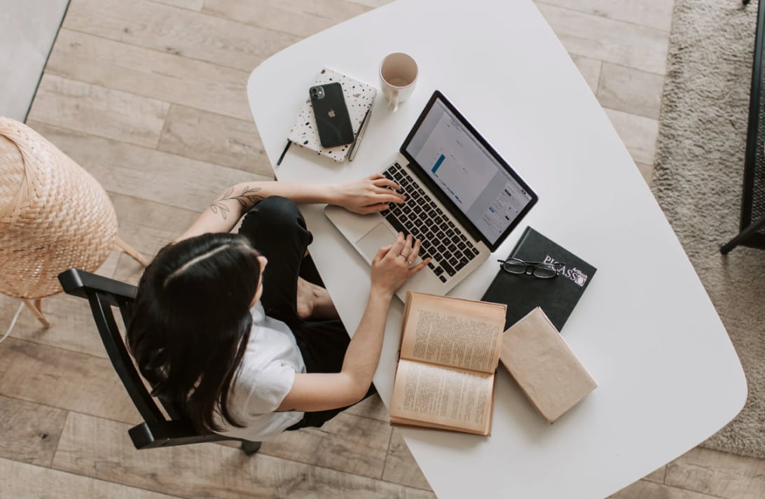 een meisje werkt op een computer, naast haar liggen boeken en notitieboekjes, een telefoon en een kopje